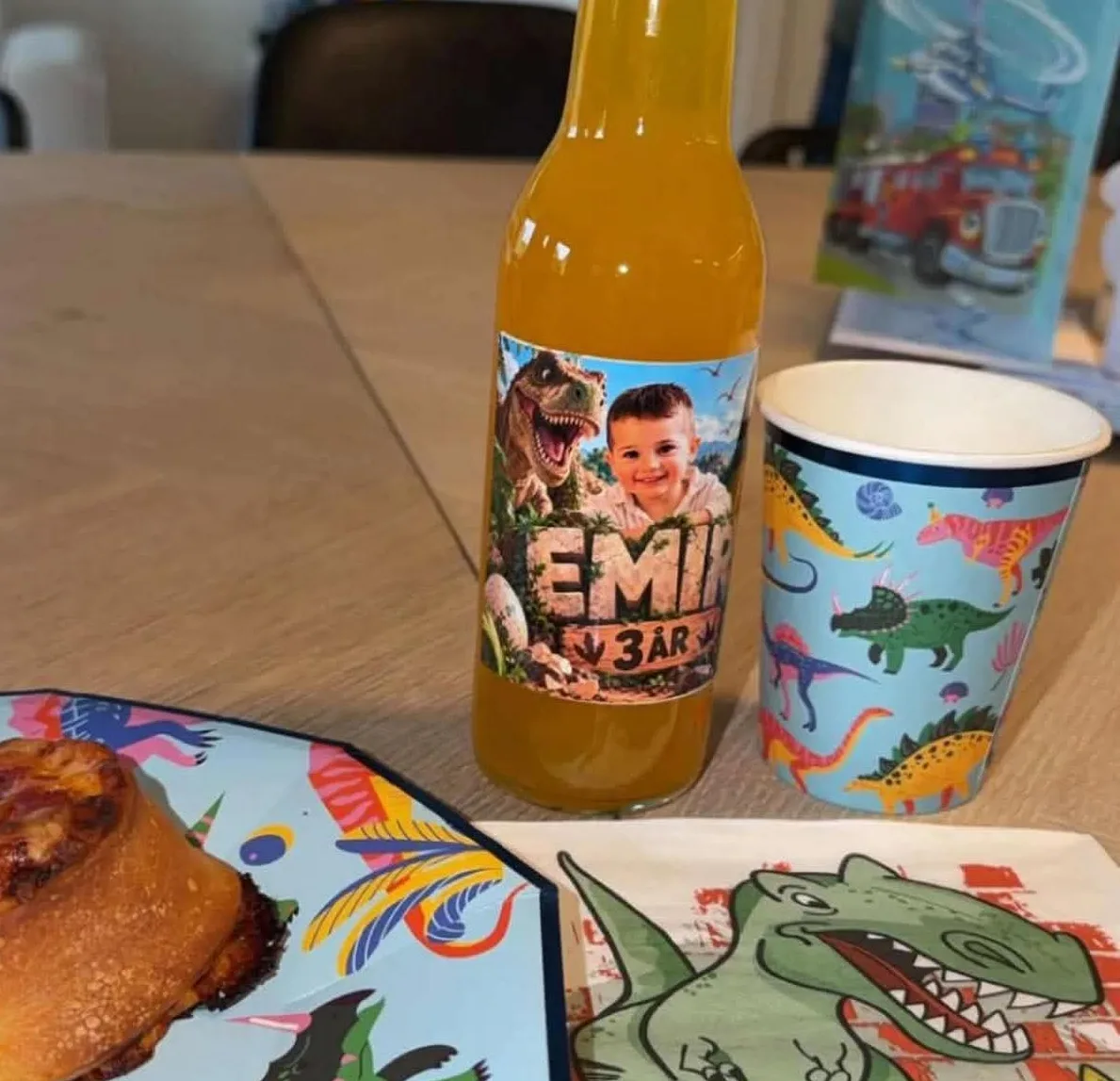 Brusflaske med dinosauretikett på bursdagsbordet, ved siden av kopp, tallerken og servietter med dinosaurer